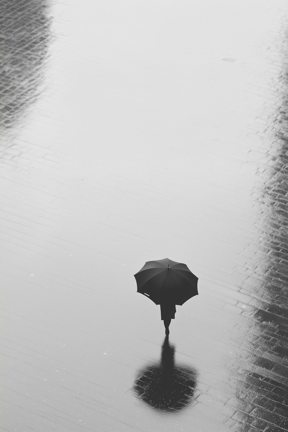 A lone figure under an umbrella reflected on wet ground, shot in black and white